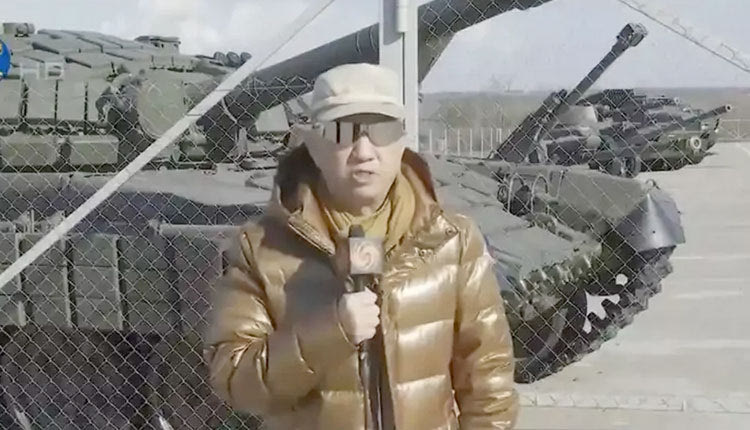 Φορώντας ένα φουσκωτό μπουφάν, κράνος και γυαλιά ηλίου, ο Κινέζος ρεπόρτερ Λου Γιουγκουάνγκ στέκεται στην άκρη ενός δρόμου και δίπλα του περνά ένα ρωσικό τανκ.