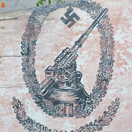 Πίνακας μεγάλων διαστάσεων που εγκατέλειψαν φεύγοντας οι Γερμανοί στο σπίτι του Εμμανουήλ Ιωάννου Καρυωτάκη (το σπίτι το είχαν επιτάξει και χρησιμοποιούνταν ως ταχυδρομείο). Ο πίνακας απεικονίζει ένα πυροβόλο της αντιαεροπορικής πυροβολαρχίας FLAK, που υπεράσπιζε το πολεμικό αεροδρόμιο Καστελλίου από τους συμμαχικούς βομβαρδισμούς 