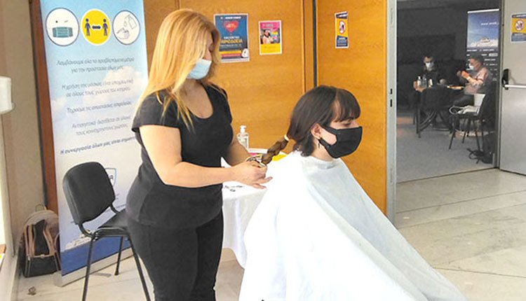 Όρθια η Διευθύντρια των κομμωτηρίων Art & Beaty, Κατερίνα Μπιτσάκη λίγο πριν κόψει από νεαρή εθελόντρια μία ακόμα κοτσίδα, η οποία προορίζεται για περούκες καρκινοπαθών του Συλλόγου "ΕΥ ΖΩ". 